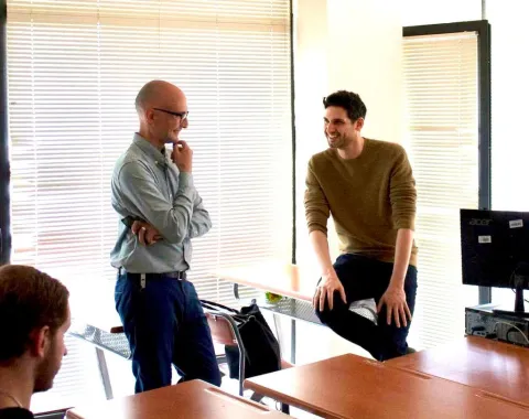 Zwei Männer in einem hellen Raum stehen und sitzen lächelnd an einem Tisch, in einem Gespräch vertieft. Im Hintergrund sind Jalousien zu sehen.