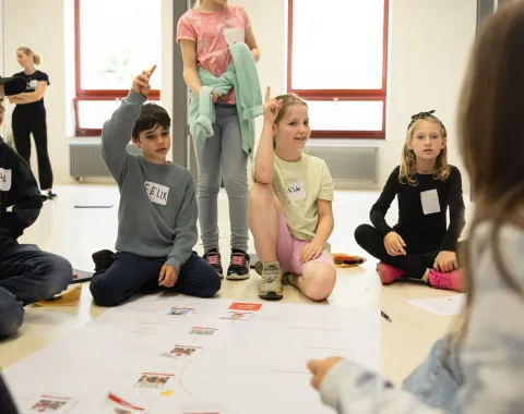 Kinder sitzen in einem Kreis auf dem Boden und nehmen an einem interaktiven Workshop teil. Einige Kinder heben die Hand, um Fragen zu beantworten. Vor ihnen liegt ein großes Papier mit Diagrammen. Die Szene findet in einem hellen Raum mit großen Fenstern statt.