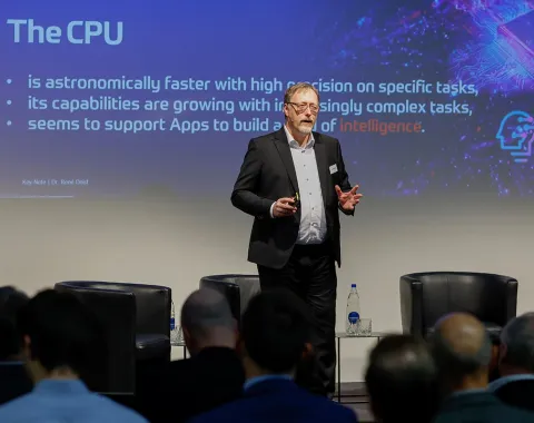 Ein Redner steht auf einer Bühne vor einem Publikum und präsentiert eine Folie über die Leistungsfähigkeit von CPUs bei einer Konferenz zur Künstlichen Intelligenz an der Universität Liechtenstein.