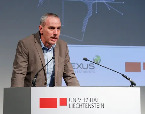 Ein Redner steht an einem Podium mit dem Logo der Universität Liechtenstein. Im Hintergrund sind Logos von PLEXUS Investments und der Universität Liechtenstein sichtbar.