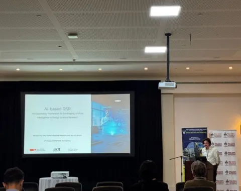 Ein Vortragender präsentiert auf einer Konferenz vor einer Leinwand mit der Folie "AI-based DSR". Im Raum sind mehrere Zuhörer zu sehen.