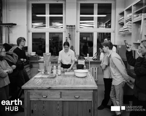 Eine Gruppe von Menschen steht in einem Workshop-Raum um einen Tisch, auf dem Materialien und Werkzeuge liegen. Eine Person in der Mitte erklärt etwas. Im Hintergrund sind Regale und Fenster zu sehen. Logos von "earth HUB" und "Universität Liechtenstein" sind eingeblendet.
