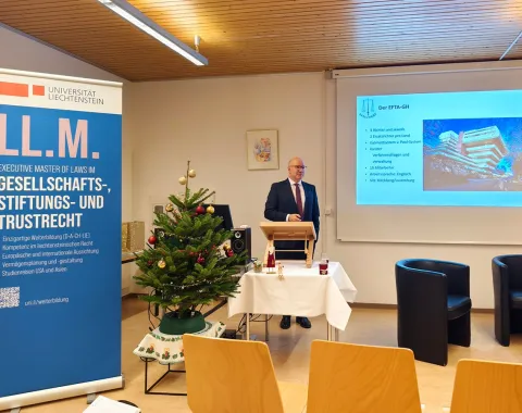 Ein Redner steht in einem Seminarraum der Universität Liechtenstein vor einem Pult. Neben ihm ist ein Banner des LL.M.-Programms für Gesellschafts-, Stiftungs- und Trustrecht zu sehen. Im Hintergrund wird eine Präsentation über den EFTA-Gerichtshof gezeigt. Ein geschmückter Weihnachtsbaum steht links im Bild.