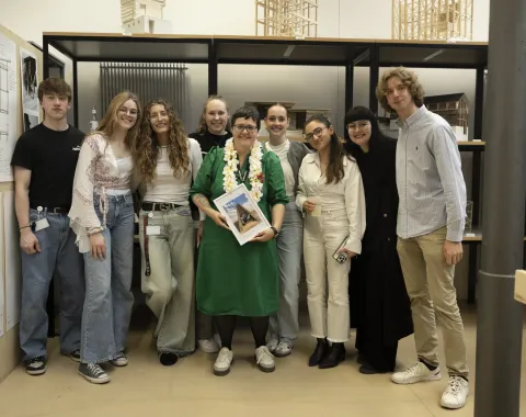 Eine Gruppe von neun Personen steht lächelnd in einem Innenraum. Eine Person in der Mitte hält ein Buch und trägt eine grüne Kleidung mit einer Blumenkette. Im Hintergrund sind architektonische Modelle zu sehen.