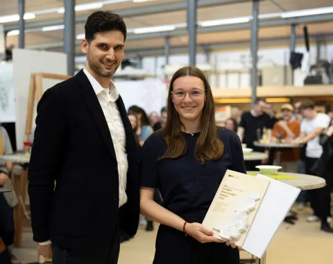 Zwei Personen stehen in einem Innenraum. Eine Person hält eine Auszeichnung oder ein Buch in den Händen. Im Hintergrund sind weitere Menschen und eine helle, moderne Umgebung zu sehen.