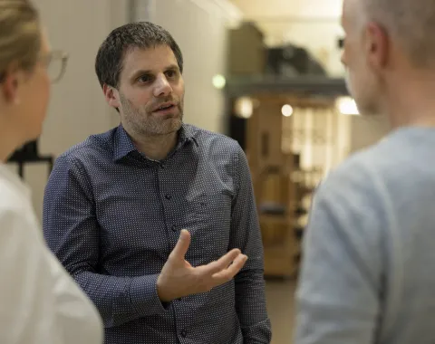 Ein Mann in einem karierten Hemd spricht gestikulierend mit zwei anderen Personen in einem Innenraum. Die Szene wirkt professionell und konzentriert.