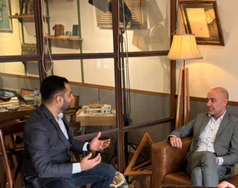 Zwei Männer in einem Gespräch in einem gemütlichen Raum mit Glaswänden und Holzmöbeln. Einer sitzt auf einem Ledersessel, der andere gestikuliert während des Gesprächs. Im Hintergrund sind Bücher und Dekorationsgegenstände zu sehen.