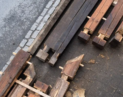 Holzbalken und Bauteile liegen auf einem gepflasterten Boden neben einem Bordstein. Die Materialien sind teilweise bearbeitet und bereit für den Einsatz in einem Bauprojekt. Das Bild zeigt den Fokus auf nachhaltige Baupraktiken und Wiederverwendung von Materialien.