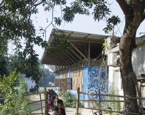 Eine Gruppe von Kindern geht einen Weg entlang, der zu einem nachhaltig gebauten Gebäude mit blauen Wänden und einem großen Dach führt. Im Vordergrund sind Bäume und ein Zaun zu sehen. Die Szene vermittelt eine ländliche, sonnige Atmosphäre.