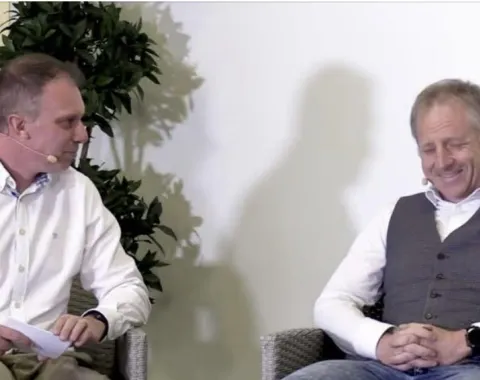 Zwei Männer sitzen auf einer Bühne und unterhalten sich. Beide tragen Mikrofone. Links sitzt ein Mann in einem weißen Hemd, rechts ein Mann in einem grauen Pullunder. Im Hintergrund ist eine Pflanze zu sehen.