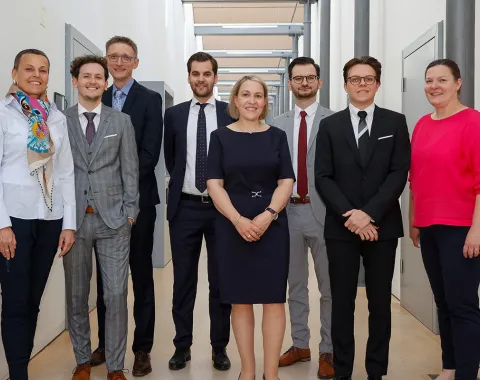 Eine Gruppe von acht Personen steht in einem Flur der Universität Liechtenstein. Sie tragen formelle Kleidung und lächeln in die Kamera. Die Umgebung ist modern und hell gestaltet.