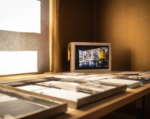 Ein Tisch mit Architekturmodellen und Dokumenten, im Hintergrund ein Bildschirm mit einer Baustellenszene.