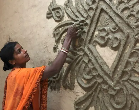 Eine Frau in einem orangefarbenen Sari verziert eine Wand mit traditionellen Tonmustern. Sie arbeitet konzentriert an einem kunstvollen Relief.