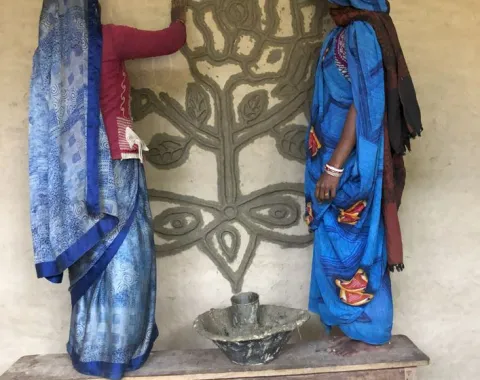 Zwei Frauen in traditionellen indischen Kleidern verzieren eine Wand mit einem kunstvollen Blumenmuster. Sie stehen auf einer Holzbank und arbeiten gemeinsam an der Gestaltung.