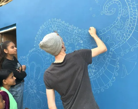 Eine Person zeichnet ein detailliertes Muster auf eine blaue Wand, während drei Kinder zuschauen. Die Szene zeigt gemeinschaftliches Engagement beim Bau einer Schule in Indien.