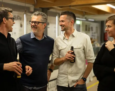 Vier Personen stehen in einem Innenraum und unterhalten sich fröhlich. Zwei von ihnen halten Getränke in der Hand. Im Hintergrund sind Ausstellungstafeln und eine Holzkonstruktion zu sehen. Die Atmosphäre wirkt entspannt und gesellig.