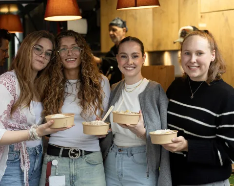Vier lächelnde Personen halten Schalen mit Essen in einem gemütlichen Innenraum. Im Hintergrund ist eine Küche zu sehen. Die Szene vermittelt eine freundliche und einladende Atmosphäre.