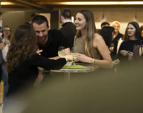 Eine Gruppe von Menschen unterhält sich fröhlich bei einem Stehempfang. Zwei Personen halten Gläser mit Getränken in der Hand. Im Hintergrund sind weitere Gäste zu sehen. Die Stimmung wirkt entspannt und gesellig.