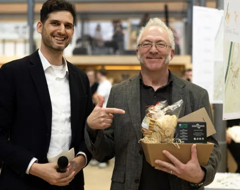 Zwei Männer stehen lächelnd in einem Innenraum. Der Mann links hält ein Mikrofon, während der Mann rechts auf einen Geschenkkorb zeigt, den er in der Hand hält. Im Hintergrund sind unscharf weitere Personen und eine moderne Architektur zu erkennen.