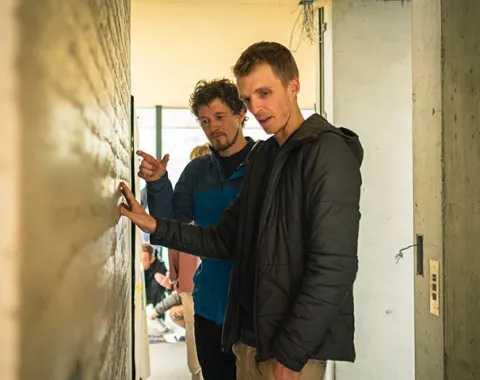 Zwei Männer betrachten und diskutieren eine Wand aus Stampflehm in einem Gebäude. Sie tragen Freizeitkleidung und befinden sich in einem hellen Raum.
