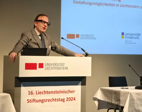 Ein Redner steht am Podium des 16. Liechtensteinischen Stiftungsrechtstags 2024 an der Universität Liechtenstein. Im Hintergrund sind Logos der Universität Liechtenstein und der Universität Innsbruck zu sehen.