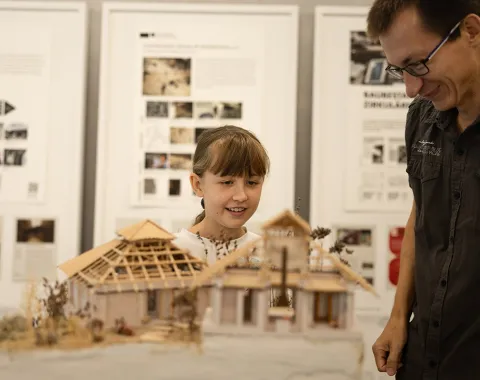 Ein Kind und ein Erwachsener betrachten ein detailliertes Architekturmodell eines Gebäudes. Im Hintergrund sind Informationsplakate zu sehen.