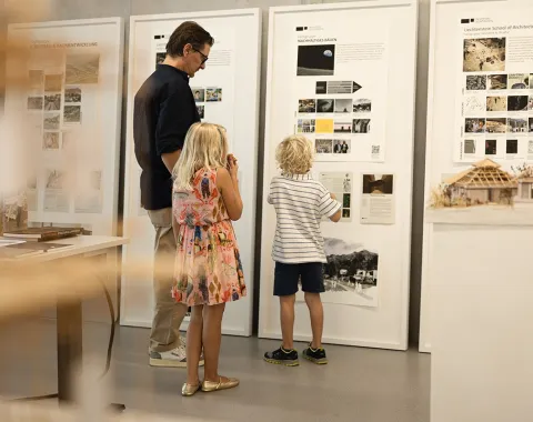 Ein Erwachsener und zwei Kinder betrachten Informationswände mit Architekturprojekten in einem Ausstellungsraum der Universität Liechtenstein.