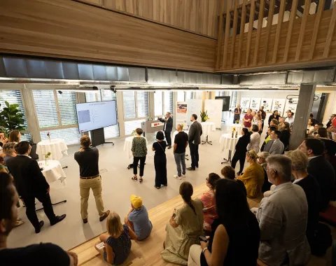 Eine Gruppe von Menschen versammelt sich in einem modernen, hellen Raum mit großen Fenstern. Im Vordergrund sitzen einige Personen auf Holzstufen. Im Hintergrund hält eine Person eine Präsentation vor einem Bildschirm. Der Raum ist mit Pflanzen und Stehtischen dekoriert.