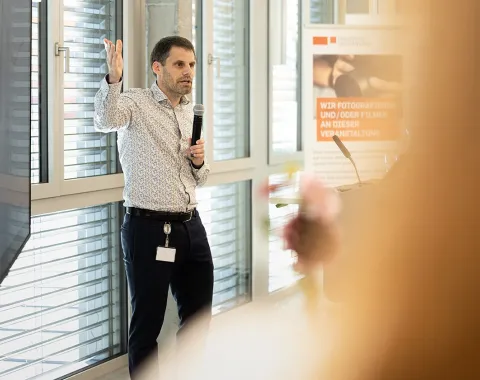 Eine Person hält eine Präsentation in einem modernen Raum mit großen Fenstern. Sie spricht in ein Mikrofon und gestikuliert mit der Hand. Im Hintergrund ist ein Plakat mit Informationen sichtbar.