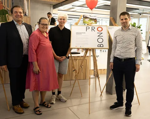 Vier Personen stehen in einem modernen Raum neben einer Ausstellungstafel mit der Aufschrift "Pro Bono". Im Hintergrund sind weitere Ausstellungstafeln und ein roter Luftballon zu sehen. Die Szene wirkt freundlich und einladend.