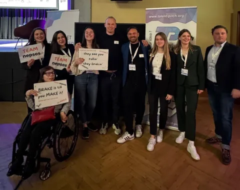 Gruppe von neun Personen posiert lächelnd bei einem Event, einige halten Schilder mit motivierenden Botschaften. Im Hintergrund sind Banner mit der Aufschrift "Talent Pitch" zu sehen. Eine Person sitzt im Rollstuhl.
