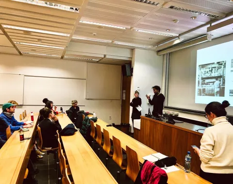 Ein Vortrag in einem Hörsaal: Zwei Personen präsentieren vor einer Gruppe von Studierenden. Auf der Leinwand ist eine Projektion zu sehen. Die Studierenden sitzen an Holztischen und hören aufmerksam zu.