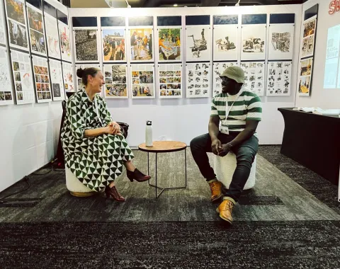 Zwei Personen sitzen in einer Ausstellung vor einer Wand mit zahlreichen Bildern und Texten. Sie unterhalten sich entspannt auf Hockern. Die Ausstellung thematisiert urbanes Design und soziale Realitäten in Afrika.