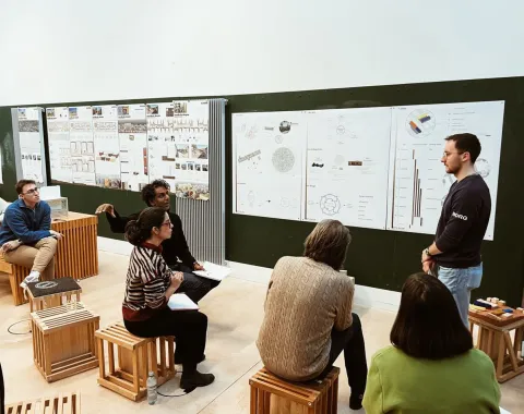 Eine Gruppe von Studierenden sitzt auf Hockern in einem Seminarraum und diskutiert vor einer Wand mit Architekturplänen und Diagrammen. Ein Präsentierender steht rechts und erklärt die Inhalte.