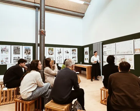 Eine Gruppe von Studierenden sitzt auf Holzhockern in einem Seminarraum und hört einer Person zu, die vor Präsentationspostern an der Wand steht. Die Atmosphäre ist konzentriert und akademisch.