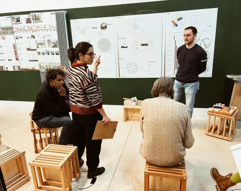 Eine Gruppe von Personen diskutiert vor einer Wand mit Architekturplänen. Sie sitzen auf Holzhockern in einem Seminarraum. Eine Frau steht und gestikuliert, während sie auf die Pläne zeigt.