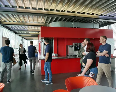 Eine Gruppe von Menschen steht in einem modernen Raum mit roten Wänden und einer Kaffeestation, möglicherweise während eines Treffens an der Universität Liechtenstein.