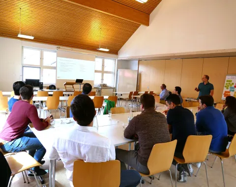Eine Gruppe von Menschen sitzt in einem Seminarraum der Universität Liechtenstein und hört einer Präsentation zu. Ein Mann steht vorne und spricht, während eine Präsentation auf eine Leinwand projiziert wird. Die Teilnehmer sitzen an Tischen und sind aufmerksam.