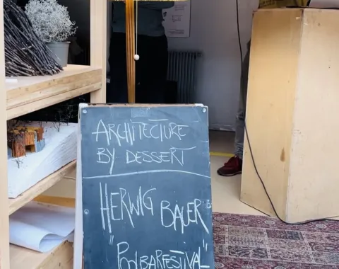 Ein Mann spricht in ein Mikrofon neben einem Rednerpult. Im Vordergrund steht eine Tafel mit der Aufschrift „Architecture by Dessert, Herwig Bauer, Poolbarfestival“. Links sind ein Regal und eine Stehlampe zu sehen.