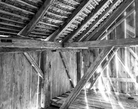 Schwarz-Weiß-Foto eines alten Dachbodens mit sichtbaren Holzbalken und einfallendem Licht durch die Dachlücken. Die Struktur zeigt traditionelle Bauweise und vermittelt eine rustikale Atmosphäre.