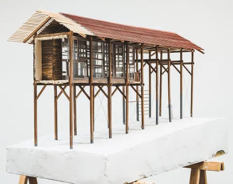 Ein Architekturmodell eines Strickbaus auf Stelzen, das auf einem weißen Sockel präsentiert wird. Das Modell zeigt eine lineare Struktur mit einem Satteldach und großen Fenstern, die traditionelle Bauweise und moderne Elemente kombinieren.