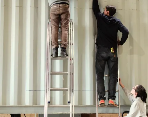 Drei Personen arbeiten an einem großen, weißen Container. Zwei stehen auf Leitern und befestigen etwas an der Wand, während die dritte Person unten steht und assistiert.