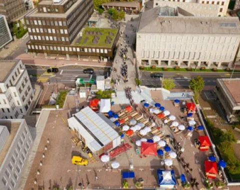Luftaufnahme eines städtischen Platzes mit dem Base Camp der Universität Liechtenstein. Zu sehen sind modulare Strukturen, bunte Zelte und Menschen, die sich auf dem Platz bewegen. Gebäude umgeben den Platz, und eine Straße verläuft im Hintergrund.