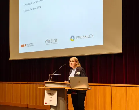 Eine Frau steht an einem Rednerpult vor einer Leinwand mit der Aufschrift „Herzlich willkommen! Compliance-Day 2024, Universität Liechtenstein, Schaan, 14. Mai 2024“. Im Hintergrund sind Logos von Sponsoren zu sehen.