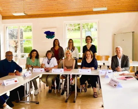Eine Gruppe von zwölf Personen sitzt an einem U-förmigen Tisch in einem hellen Raum. Sie sind Teil eines internationalen Besuchsprogramms an der Universität Liechtenstein. Im Hintergrund sind große Fenster und eine Holzbalkendecke zu sehen.