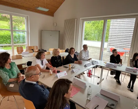 Eine Gruppe von Menschen sitzt an einem Konferenztisch in einem hellen Raum mit großen Fenstern. Sie sind in eine Diskussion vertieft. Auf dem Tisch liegen Namensschilder und Dokumente. Im Hintergrund ist eine grüne Landschaft zu sehen.