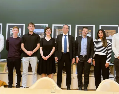 Eine Gruppe von sieben Personen, darunter Studierende und zwei Männer in Anzügen, steht lächelnd vor einer Wand mit Bildern. Sie befinden sich in einem Raum der Universität Liechtenstein.