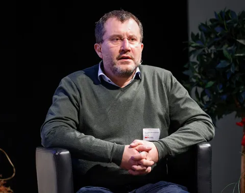 Eine Person sitzt auf einem schwarzen Sessel und hält die Hände gefaltet. Im Hintergrund sind Pflanzen zu sehen.