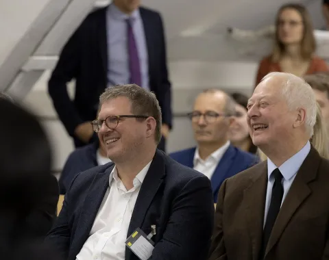 Teilnehmer einer Podiumsdiskussion an der Universität Liechtenstein, lächelnd und aufmerksam, in einem gut besuchten Veranstaltungsraum.