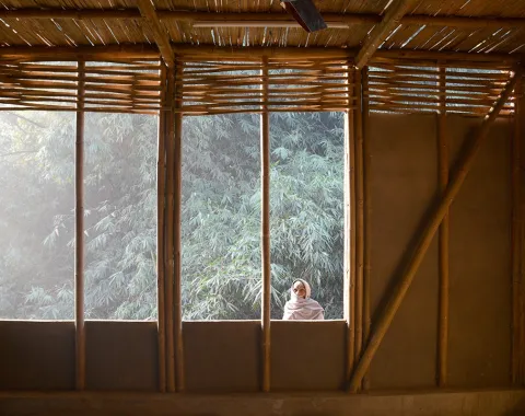 Innenansicht eines Gebäudes mit Wänden aus Lehm und Bambus, durch ein großes Fenster fällt Sonnenlicht auf eine Person, die draußen steht. Im Hintergrund sind Bäume zu sehen.